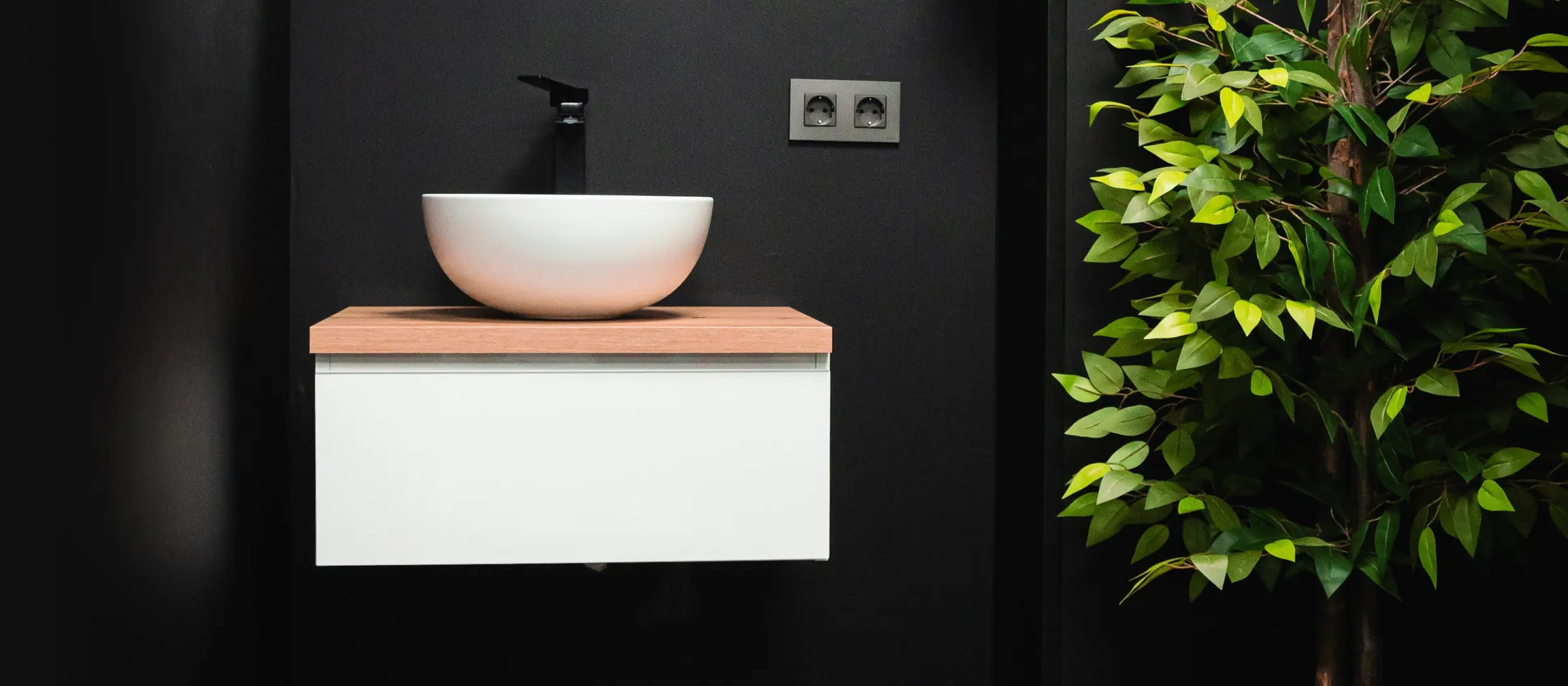 Aseo del local reformado: lavabo sobre mueble volado en pared negra, junto a una planta